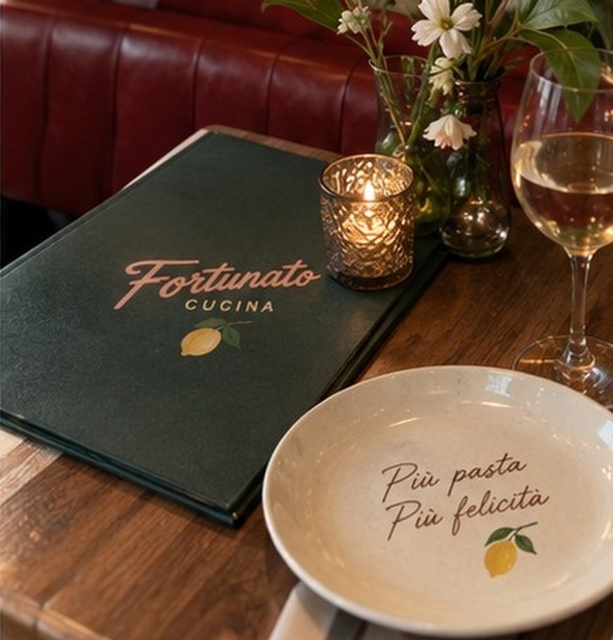 A Fortunato Cucina menu, candle and wine glass on a timber table by the red banquette